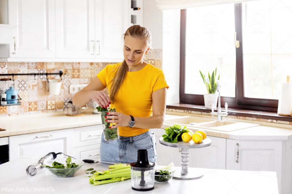Vrouw in een lichte keuken bereidt een groene smoothie met spinazie, bleekselderij en citroen.
